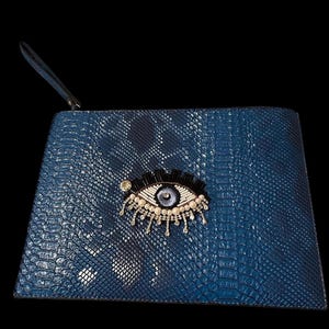May include: A blue clutch bag with a snakeskin effect, adorned with an eye motif. The eye is embellished with black, white, and silver beads, and a dangling fringe. It has a wrist strap.