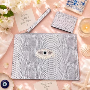 Op de afbeelding: Een zilveren clutch met slangenleerpatroon, polsband en bijpassende kaarthouder. De clutch is voorzien van een centrale kraalversiering in de vorm van een oog. Daarnaast liggen een kaart met Island Nights & Good Vibes en een Mykonos-onderzetter.