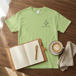 Könnte beinhalten: Ein hellgrünes T-Shirt mit einer kleinen schwarzen Grafik einer Person, die einen Laptop benutzt, und dem Text "Eat, Sleep, CAD, Repeat". Ein braunes Leder-Notizbuch, ein goldener Stift, eine Kaffeetasse und eine Serviette liegen ebenfalls auf der Holzoberfläche.