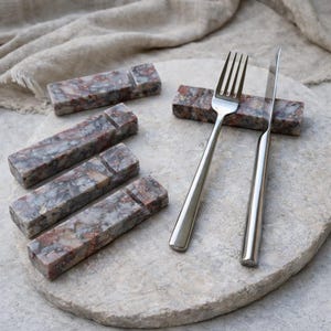 May include: A set of six rectangular marble knife rests with a notch for cutlery. The rests are gray, red, and white. A stainless steel fork and knife rest on one of the marble rests, displayed on a stone serving platter.
