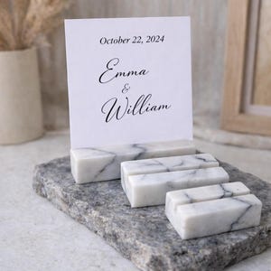 Pode incluir: Um cartão branco com o texto "October 22, 2024" e os nomes "Emma & William" é segurado por porta-cartões de mármore. Os suportes estão sobre uma base de pedra cinza. A cena é bem iluminada.