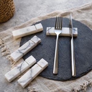 May include: A set of white marble utensil rests with gray veining. A fork and knife rest on a black slate plate. Additional rests are scattered around the plate on a neutral-colored linen cloth. The rests are rectangular with a groove.