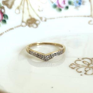 May include: A gold V-shaped ring featuring a row of small, clear, sparkling stones set along the chevron-style band. The ring has a polished metallic finish and a pointed centre design that creates a distinct angular silhouette on the finger.