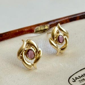 May include: A pair of gold stud earrings featuring a polished, swirling metal design that frames a central, oval-shaped, deep pink gemstone. The earrings have a smooth, reflective finish and are presented in a light-coloured, fabric-lined display case.