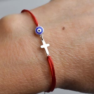 May include: Red string bracelet featuring a silver cross charm and a blue and white evil eye charm. The bracelet is worn on a wrist, showcasing the vibrant red cord and the detailed charms.