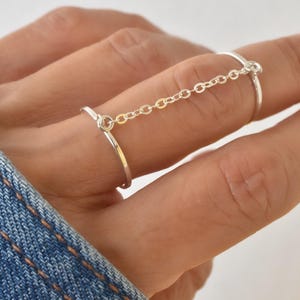 May include: A silver ring set connected by a delicate chain. The ring set is worn on two fingers, with a simple, elegant design. The jewelry is displayed against a denim background.