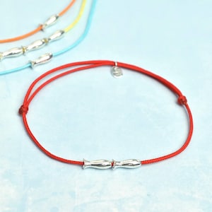 May include: A red bracelet with a silver fish charm and a small silver tag. Several other bracelets with silver fish charms are visible, featuring blue, yellow, and orange cords. The bracelets are on a light blue background.