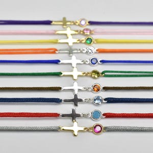 May include: A collection of colorful cord bracelets with cross pendants. Each bracelet features a different colored cord, including purple, pink, yellow, orange, blue, green, brown, navy, red, and gray. The crosses are gold or silver, with a gemstone accent.