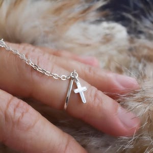 May include: A silver ring set with a chain connecting two rings. One ring is on the index finger, and the other is on the middle finger. A small cross charm hangs from the chain. The ring is shown against a blurred background.