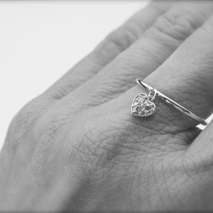 May include: A silver ring with a dangling heart charm. The heart is covered in small, sparkling stones.