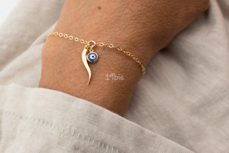 May include: Gold chain bracelet with a blue evil eye charm and a gold horn charm. The bracelet is on a person's wrist.