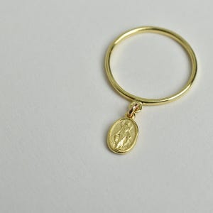 Peut inclure: Une bague en or avec un petit pendentif ovale repr&eacute;sentant une figure religieuse. La bague est simple et d&eacute;licate.