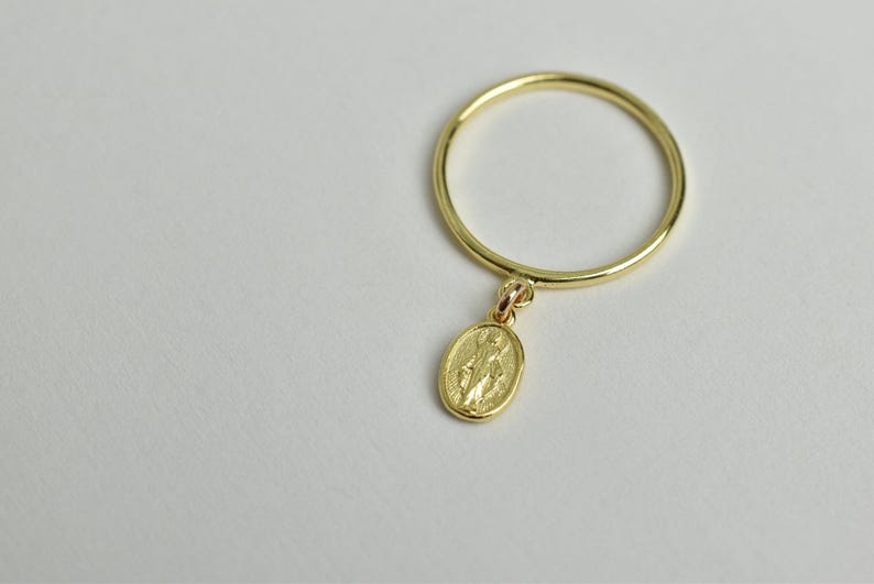 Peut inclure: Une bague en or avec un petit pendentif ovale repr&eacute;sentant une figure religieuse. La bague est simple et d&eacute;licate.