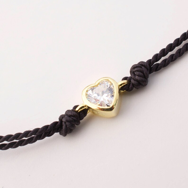 May include: A black cord bracelet with a gold heart-shaped pendant featuring a clear cubic zirconia stone.