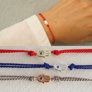 May include: Three red, blue, and gray string bracelets with a gold, silver, and rose gold hamsa hand charm with a blue evil eye. The charms are all the same size and shape.