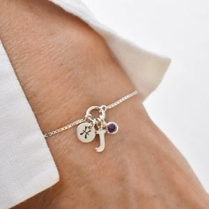 May include: Silver charm bracelet with a delicate chain. The bracelet features a round charm with a zodiac symbol, a letter "J" charm, and a small purple gemstone. The bracelet is worn on a wrist.
