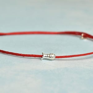 May include: A red cord bracelet featuring a small silver fish charm. The bracelet has a simple design with a knot fastening. The fish charm is detailed, adding a playful element. This bracelet is a piece of jewellery.
