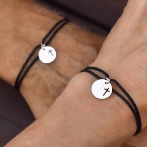 May include: Two black cord bracelets, each featuring a silver-colored circular charm with a cross design. The bracelets are worn on wrists, showcasing the simple, elegant design. The background is blurred.