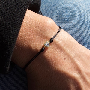 May include: A black string bracelet with a small gold heart charm featuring a clear crystal.