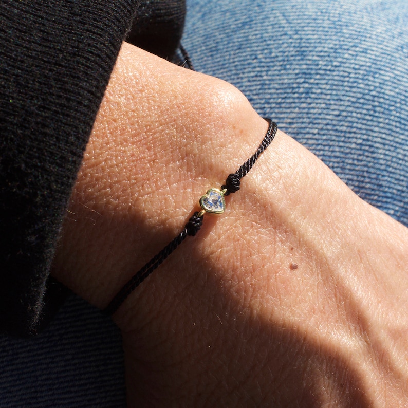 May include: A black string bracelet with a small gold heart charm featuring a clear crystal.