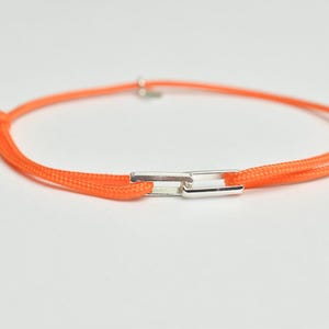 May include: An orange bracelet with a silver clasp. The bracelet is made of a thin, woven cord and features a rectangular silver clasp in the center. The cord is knotted on each end.