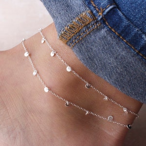 May include: Two silver anklets with delicate chains and small round charms dangling from them. The anklets are layered on a person's ankle.