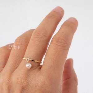 Puede incluir: Un anillo de oro con una pequeña perla colgando. El anillo se lleva en un dedo.