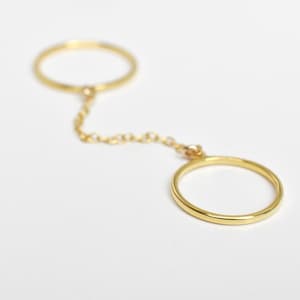 May include: A gold-colored ring with two circular bands connected by a delicate chain. The rings are simple in design, with a smooth, polished finish. The jewelry piece is set against a plain white background, highlighting its elegant simplicity.