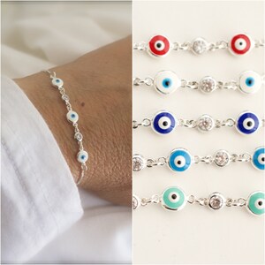 Puede incluir: Un brazalete de cadena de plata con cinco amuletos de ojo azul maligno y detalles de cristal transparente. El brazalete se muestra en la mu&ntilde;eca de una persona.