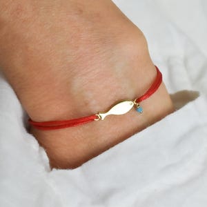 Puede incluir: Una pulsera de cordón rojo con un dije de pez dorado y una pequeña cuenta turquesa. La pulsera se lleva en la muñeca, sobre un fondo blanco.
