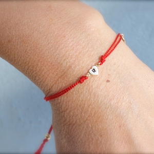 May include: A red string bracelet with a small gold heart charm featuring the letter "R" on a white background.