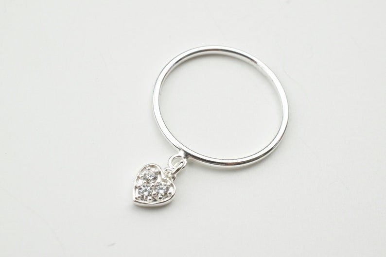 May include: A silver ring with a dangling heart charm. The heart is decorated with small clear stones.