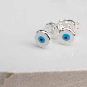 May include: A pair of silver earrings with blue evil eye charms. The earrings are small and delicate, and the evil eye charms are set in a silver circle.