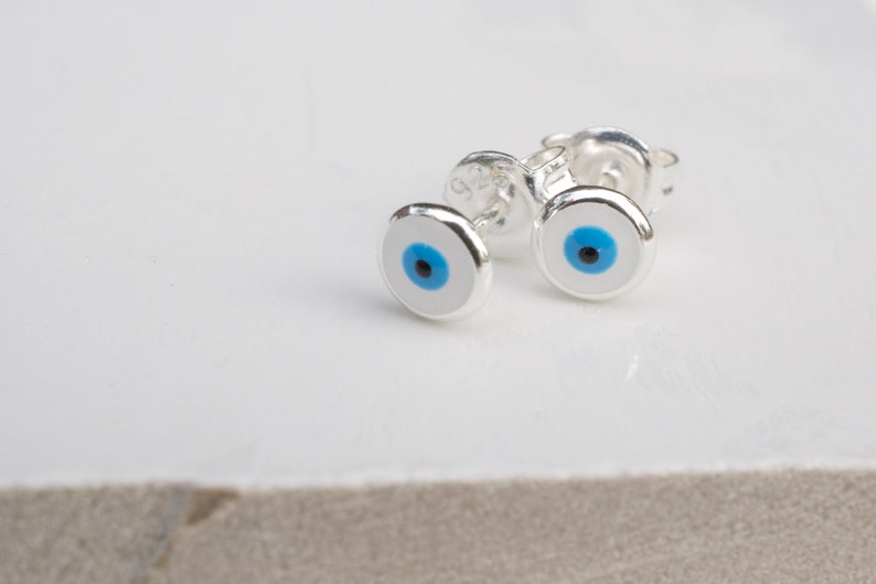 May include: A pair of silver earrings with blue evil eye charms. The earrings are small and delicate, and the evil eye charms are set in a silver circle.