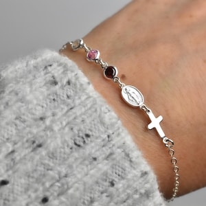 May include: A silver rosary bracelet featuring a cross, an oval pendant with a religious figure, and several colorful beads. The bracelet is worn on a wrist, with a gray sweater in the background.