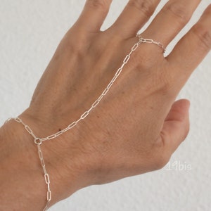 May include: A silver chain bracelet with a ring attached to the end. The bracelet is worn on the wrist and the ring is worn on the middle finger.