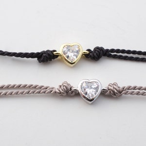 May include: Two heart-shaped bracelets with a clear crystal center. The top bracelet has a gold setting and a black cord. The bottom bracelet has a silver setting and a light brown cord.