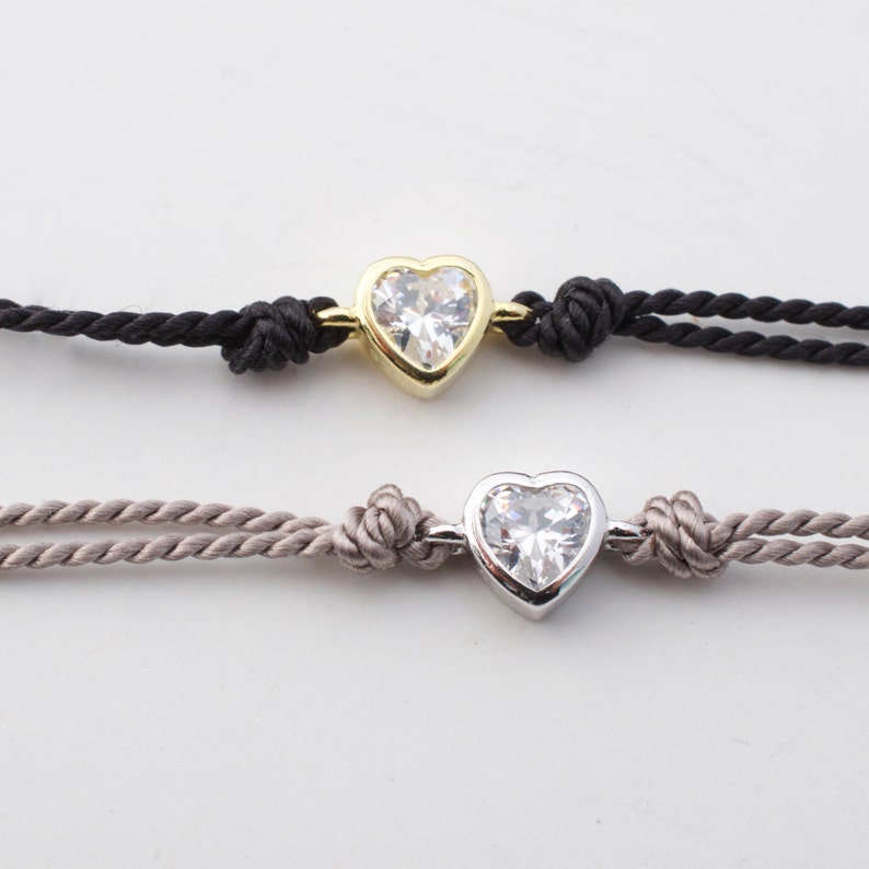 May include: Two heart-shaped bracelets with a clear crystal center. The top bracelet has a gold setting and a black cord. The bottom bracelet has a silver setting and a light brown cord.