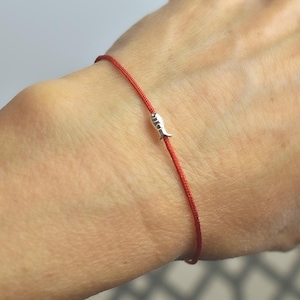 May include: A red string bracelet with a small silver fish charm. The bracelet is worn on a wrist, showcasing the simple design and the contrast between the red string and the silver charm. The jewelry is delicate and minimalist.