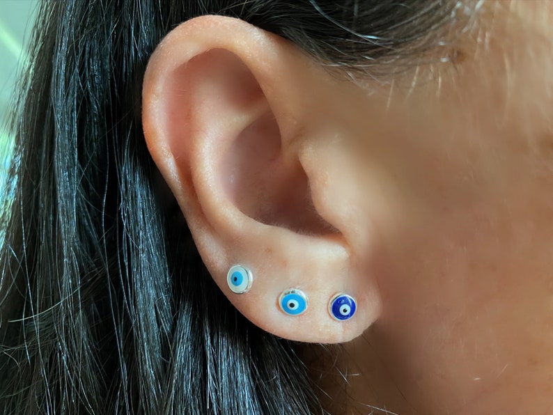 May include: Three silver evil eye earrings with blue and white enamel. The earrings are small and delicate, and they are perfect for adding a touch of style to any outfit.