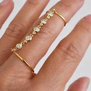 May include: A gold-colored ring with a chain of small, clear gemstones connecting two bands. The ring is worn on a finger, with one band on the base of the finger and the other on the middle knuckle. The gemstones are round and evenly spaced.