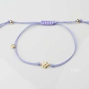 May include: A delicate lilac bracelet with a gold flower charm. The bracelet is made of a thin, braided cord and has gold beads at each end.