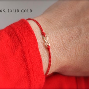 May include: A red string bracelet with a gold infinity charm. The bracelet is worn on a wrist, and the sleeve of a red shirt is visible. The text "14K SOLID GOLD" is in the upper left corner.