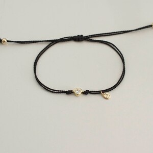 May include: A black string bracelet with a gold heart charm and a small gold tag. The charm has a clear stone in the center.