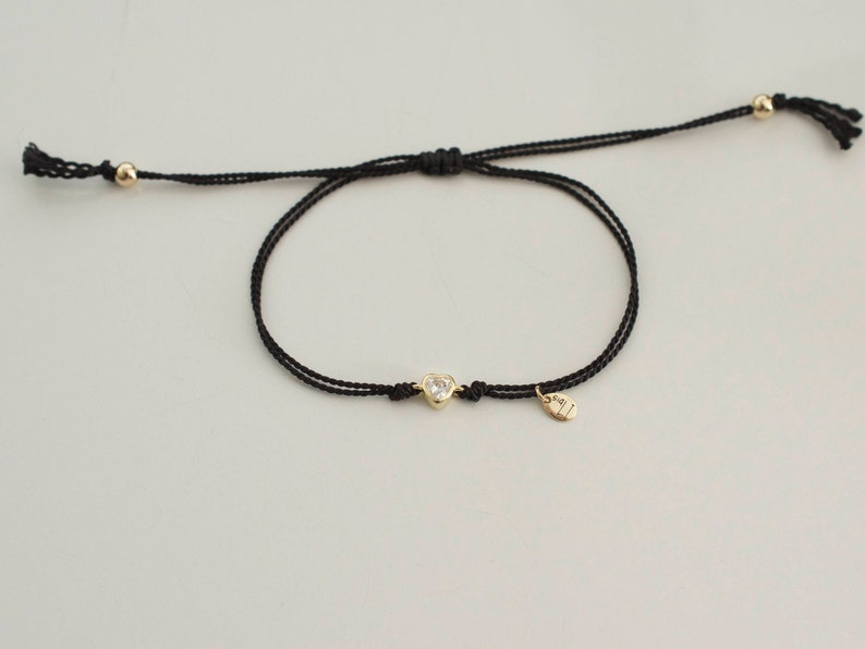 May include: A black string bracelet with a gold heart charm and a small gold tag. The charm has a clear stone in the center.