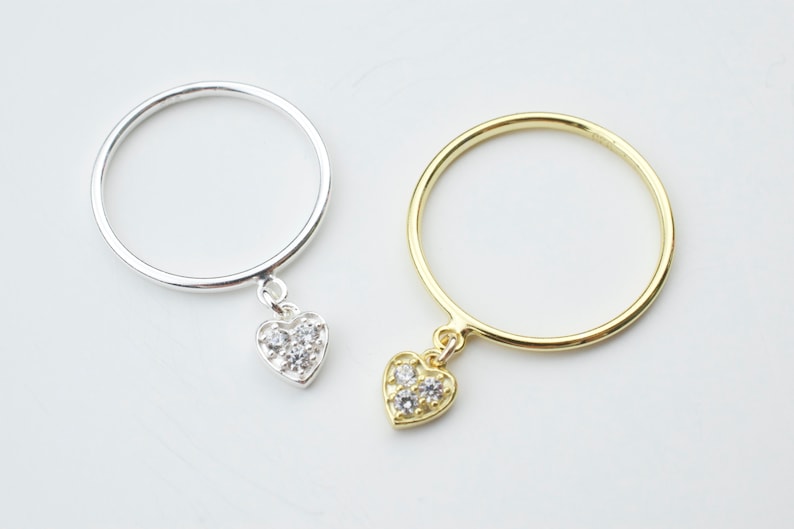 May include: Two delicate rings with a dangling heart charm. The ring on the left is silver with a clear crystal heart charm. The ring on the right is gold with a clear crystal heart charm.