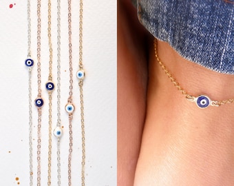 May include: A set of delicate gold chain anklets with a small blue evil eye charm on each. The anklets are shown on a white background and on a person's ankle.