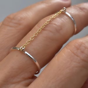 May include: A silver ring connected to a second silver ring by a delicate gold chain. The rings are worn on two fingers. The chain is made of small, linked circles. The rings have a simple, elegant design.