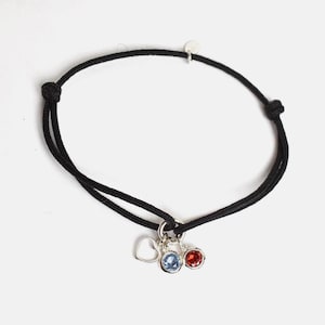 Puede incluir: Pulsera de cordón negro con un dije de corazón plateado y dos dijes de piedras preciosas, uno azul y otro rojo. La pulsera tiene nudos ajustables. Joyería.