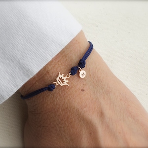 May include: A navy blue cord bracelet with a gold crown charm and a gold initial charm. The initial charm is the letter "M".
